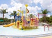 A colorful water playground with slides, sprayers, and climbing structures in a pool. Palm trees and a cloudy sky are in the background.
