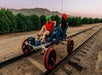 Two people wearing helmets ride a pedal-powered rail car on train tracks outdoors, with greenery and mountains in the background.