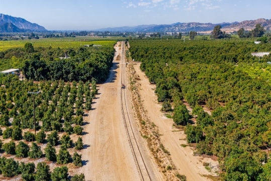 Aerial view of a railway track running through agricultural fields with trees on both sides, mountains visible in the background under a clear sky.
