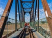 Three people wearing helmets sit on a small rail cart atop a metal railway bridge under a blue sky with scattered clouds.