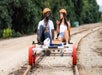 Two people wearing helmets ride a rail bike on train tracks, surrounded by greenery. The bike has red wheels and a small basket.