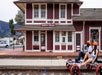 A man and woman wearing bright orange helmets on the Sunburst Railbikes pedaling past an older red and white building with several windows.