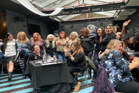 A group of women laughing and posing together inside a venue with black walls and checkered floor, some seated while others stand behind them.