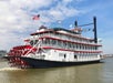 A white and red paddlewheel riverboat named "City of New Orleans" travels on a river, with multiple flags flying from its upper decks.