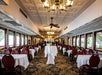 Elegant dining room with chandeliers, round tables covered in white cloths, red chairs, and large windows on both sides, set for an event or meal.