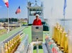 A person in a red shirt stands at the helm on the deck of a vessel, surrounded by equipment, with flags and a shoreline visible in the background.