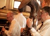 Musicians in white shirts play instruments, including a sousaphone and clarinet, during an indoor performance.