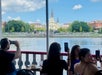 People seated on a boat deck take photos of the waterfront view of St. Louis Cathedral and Jackson Square across the river in New Orleans on a sunny day.