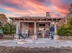 Four people pose in front of a rustic log cabin with a wooden porch, surrounded by trees, under a colorful sunset sky.