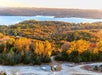 A landscape view of fall foliage covering rolling hills near a lake, with a dirt road in the foreground and water and distant shores in the background.