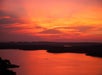 A lake at sunset with vibrant orange and pink clouds reflecting on the water, dark landmasses and hills in the distance, and a small boat on the lake.