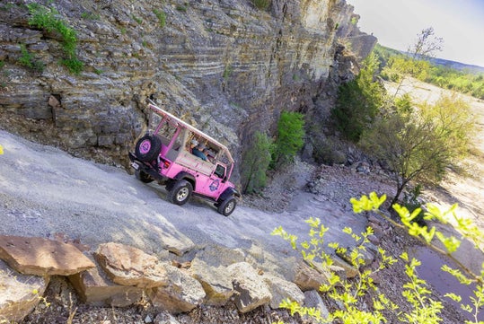 A pink open-air jeep drives up a steep, rocky trail beside a cliff, with passengers seated inside and trees visible in the background.