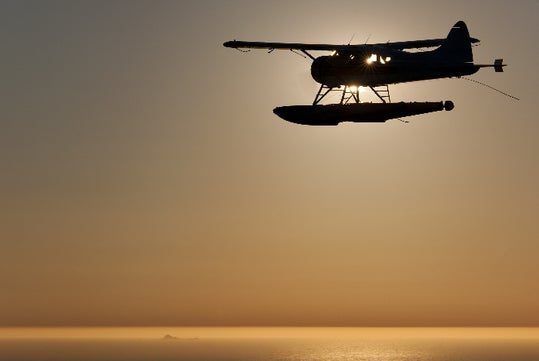 A floatplane flies above the water at sunset, silhouetted against the sky with the sun partially visible behind it.