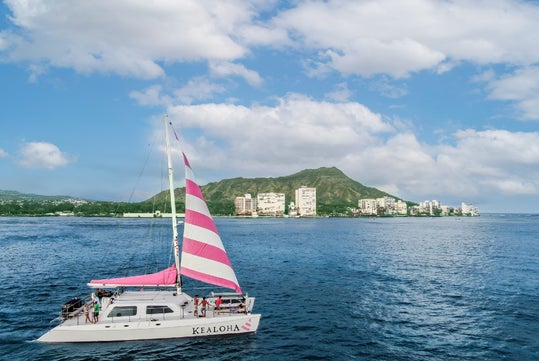 A catamaran with a pink and white sail on the ocean, with a distant view of a cityscape and a green mountain under a partly cloudy sky.