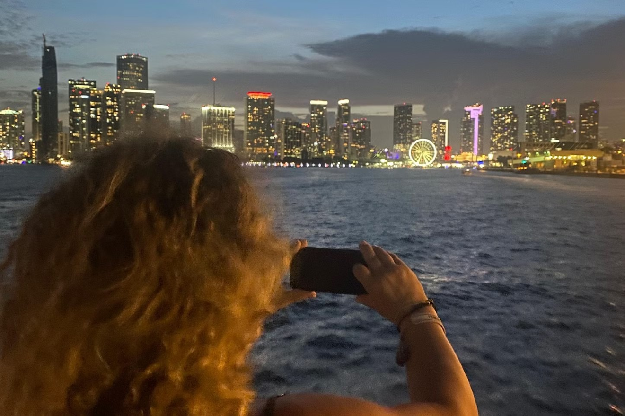 A person with curly hair takes a photo of a city skyline at night, featuring illuminated skyscrapers and a Ferris wheel across a body of water.
