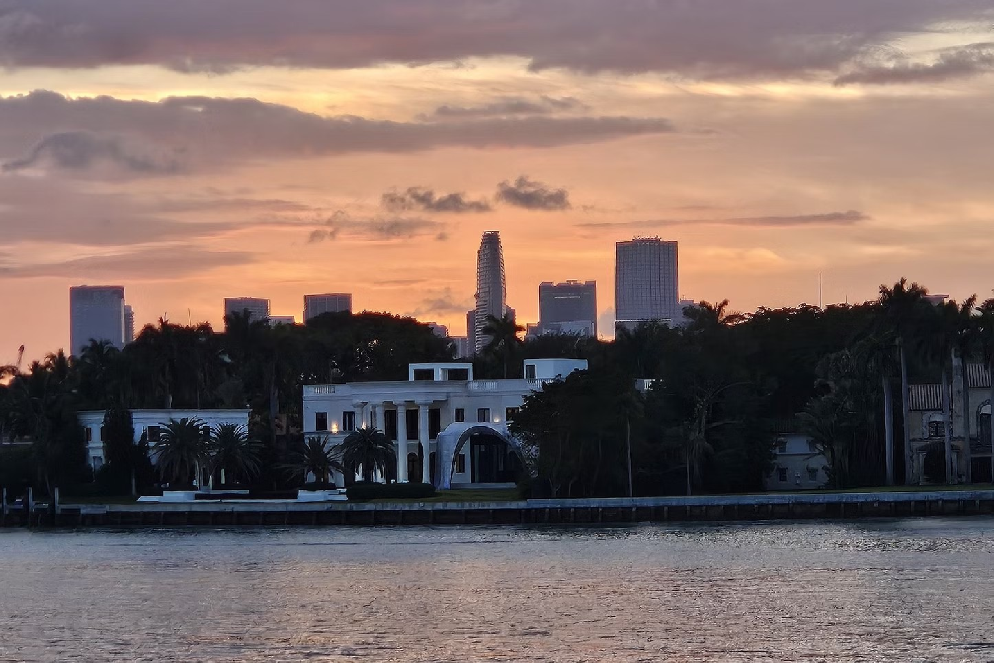 A waterfront mansion surrounded by trees with a backdrop of a city skyline at sunset.