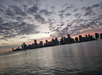 City skyline at dusk with buildings silhouetted against a cloudy sky. Water in the foreground reflects the fading light.