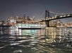 A brightly lit boat floats on the water at night with a city skyline and a suspension bridge illuminated in the background.