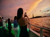 People on a boat take photos of the sunset over the water, with the Statue of Liberty visible in the distance.
