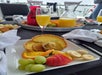 A square plate with a quiche, sliced orange, banana, grapes, and a strawberry sits on a table with juice, pastries, and a glass of mimosa on a boat.