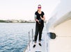 A woman wearing sunglasses holds a bottle of wine while standing on the deck of a yacht, with water and houses visible in the background.