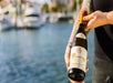 A person with tattooed arms holds a bottle of Domaine Pichot Vouvray wine outdoors near a blurred waterfront background.