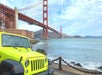 A bright yellow Jeep is parked near the waterfront with the Golden Gate Bridge and Fort Point visible in the background under a partly cloudy sky.