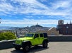 A bright green Jeep is parked on a hill overlooking a cityscape with distant buildings, a prominent tower, and a partly cloudy sky.
