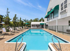Outdoor swimming pool with clear blue water, surrounded by lounge chairs, green trees, and a white building with green awnings under a clear sky.