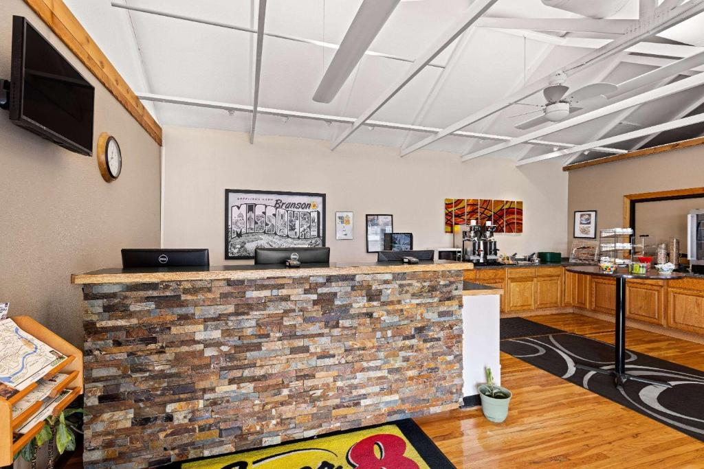 Hotel lobby with stone front desk, wall clock, artwork, TV, and brochures. Coffee station and breakfast area visible on the right with wooden cabinets and counter.