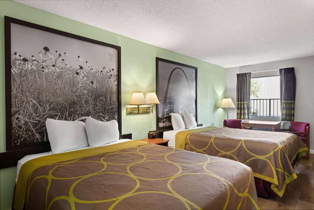 Hotel room with two double beds, green walls, large black-and-white artwork, wall lamps, window with closed curtains, and a small desk with a chair.