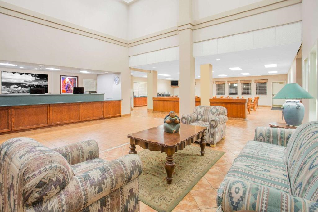 Hotel lobby with patterned couches, a wooden coffee table, and a reception desk. Decorative art and a large lamp are visible. The floor is tiled, and ceiling lights illuminate the space.