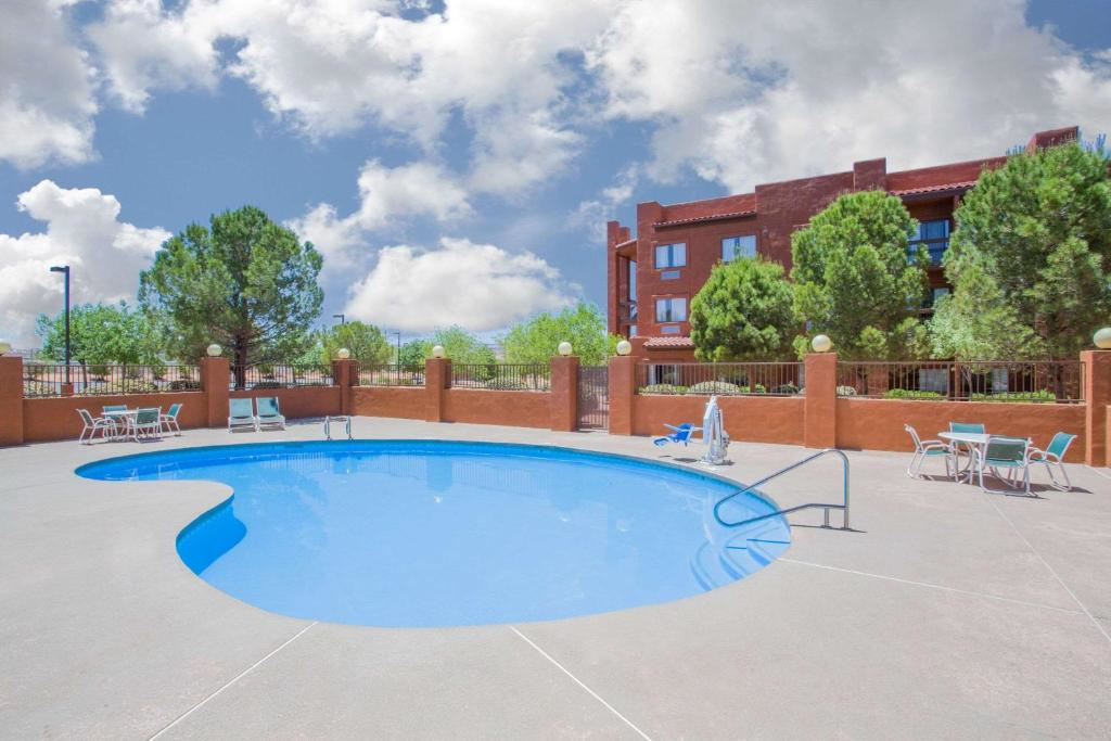 Outdoor swimming pool with blue water, surrounded by patio furniture, trees, and a red-brick building. The sky is partly cloudy.
