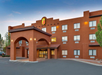 A three-story hotel with a terracotta facade, featuring an arched entrance and a Super 8 sign. The building is surrounded by a parking area and trees, set against a blue sky.