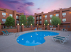 Outdoor kidney-shaped pool surrounded by lounge chairs and tables, bordered by a red stucco building with balconies, trees, and a purple-hued sky at sunset.