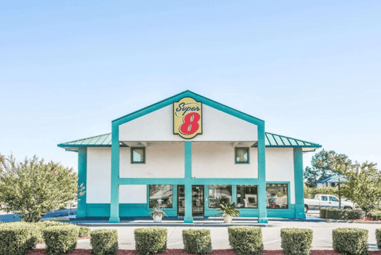 The front entrance of the Super 8 by Wyndham Valdosta/Conf Center Area, a white building with turquoise trim and roof on a bright sunny day.