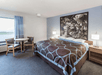 A brown and blue king bed with black and white art over it, nightstands and lamps on both sides, and a table next to a large window on the left.
