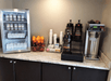 A drink station with a cooler with milk and yogurt on the left, coffee and creamers on the right, and cups, sugars, and fruit in the middle.