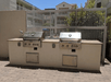 Two built-in outdoor grills are set within a masonry counter against a metal fence, with a paved surface in front.