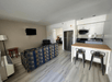 Living area with a patterned couch, armchair, wall-mounted TV, and kitchenette. Kitchenette includes a fridge, microwave, and bar seating with two stools. Wood flooring throughout.