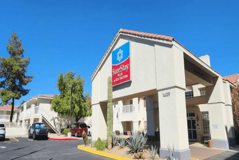 SureStay Plus Hotel exterior view with signage, parking area, and entrance. Bright day with clear blue sky and surrounding plants.