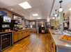 Wide view of a hotel breakfast area with wooden floors, food and beverage stations, a juice dispenser, and baskets of fruit on countertops.