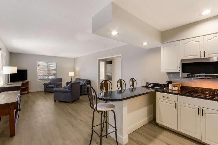 Open-concept living area with a kitchen counter and bar stools, adjacent to a seating area with a TV, armchairs, and a table. Neutral colors and wood flooring throughout.