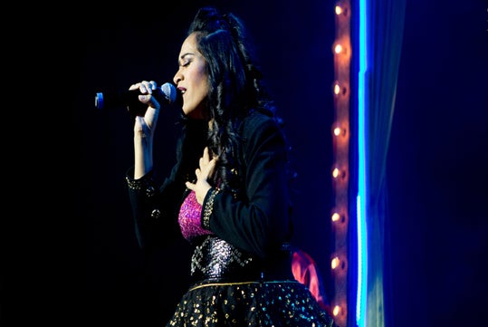 A woman performs on stage, holding a microphone with one hand and placing her other hand on her chest, wearing a black and glittery outfit.