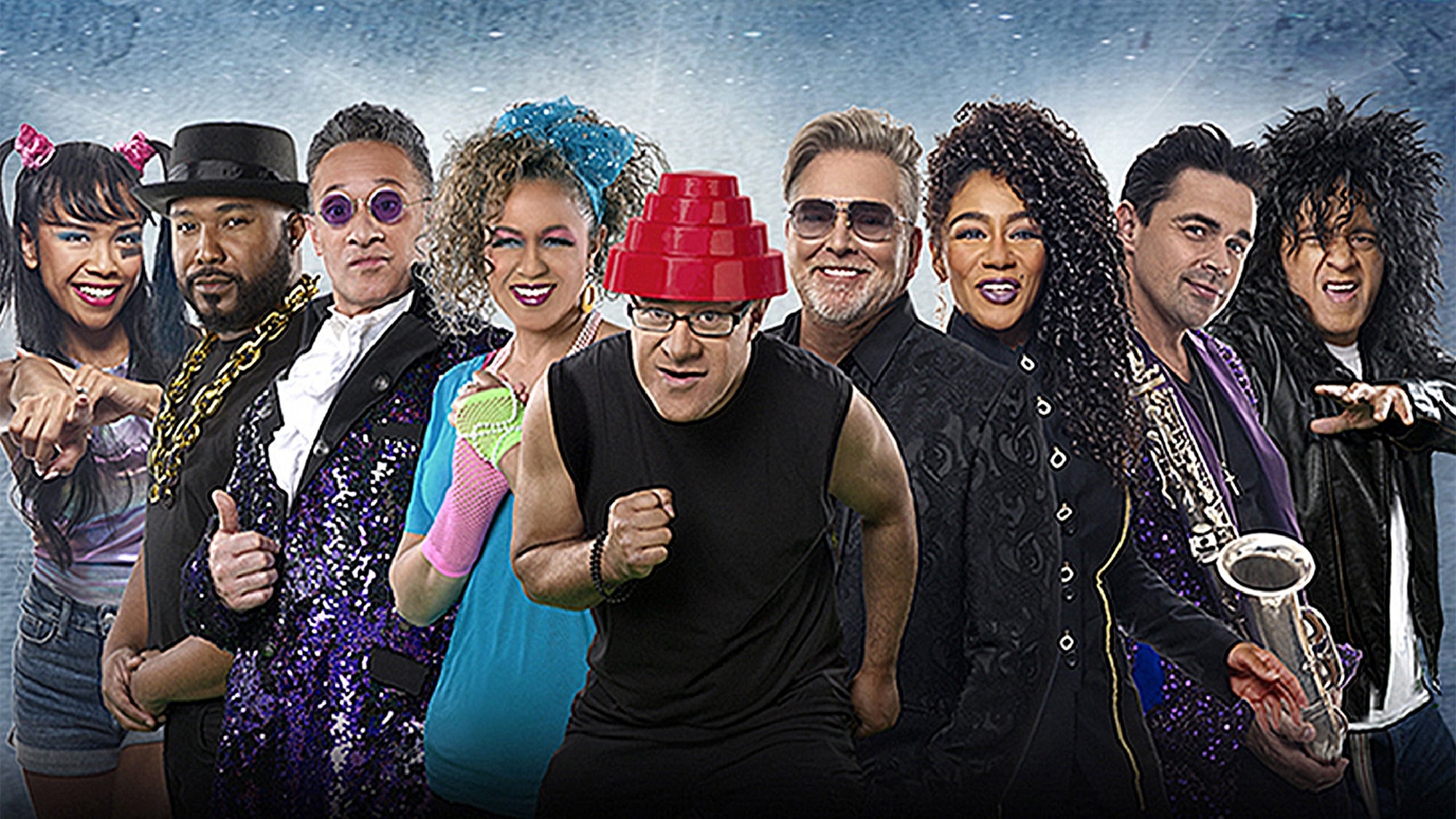 A group of nine adults dressed in colorful, retro 1980s-style costumes pose together against a blue background.