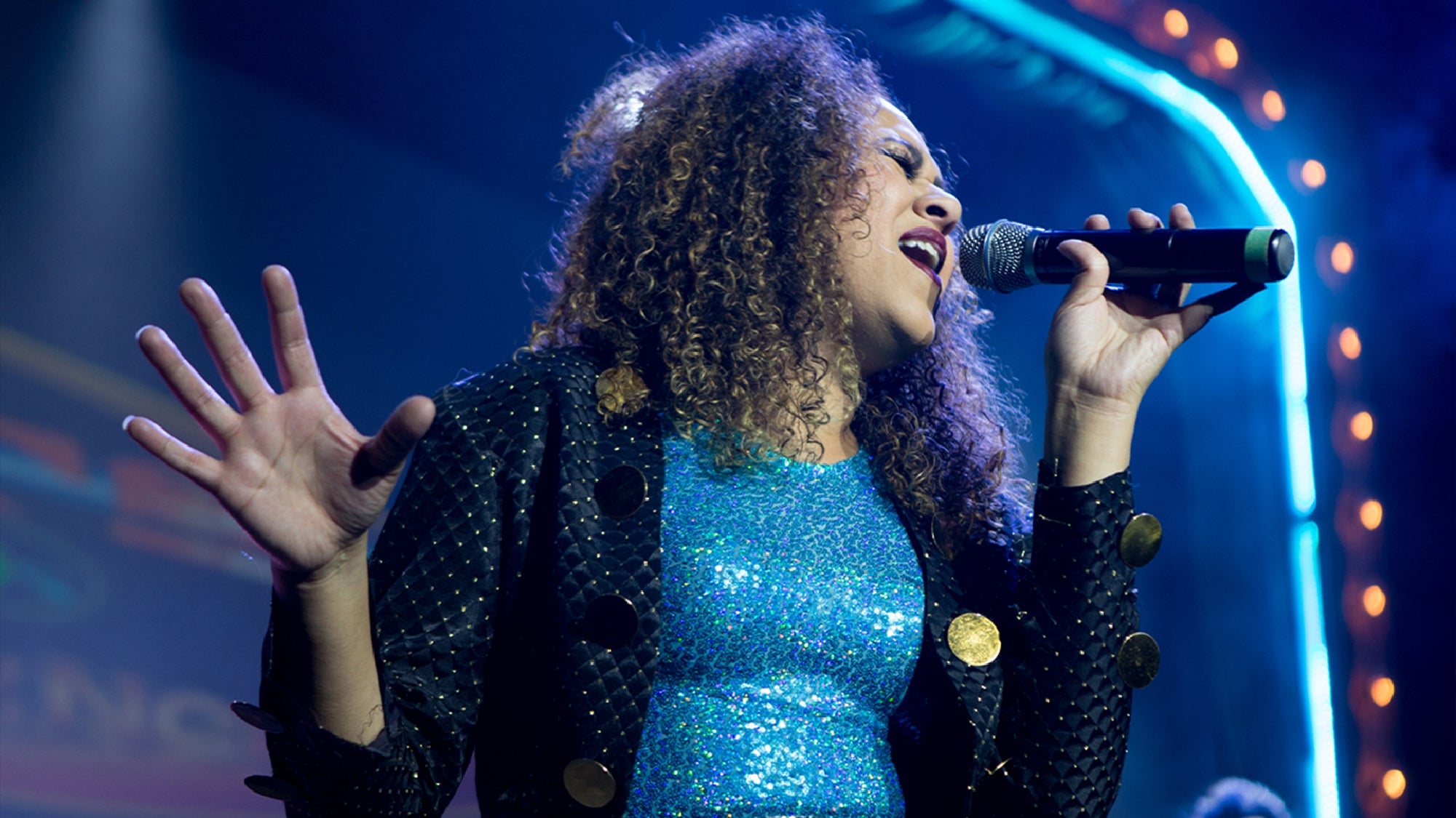 A singer with curly hair performs on stage, wearing a sparkly blue dress and holding a microphone, with stage lights in the background.