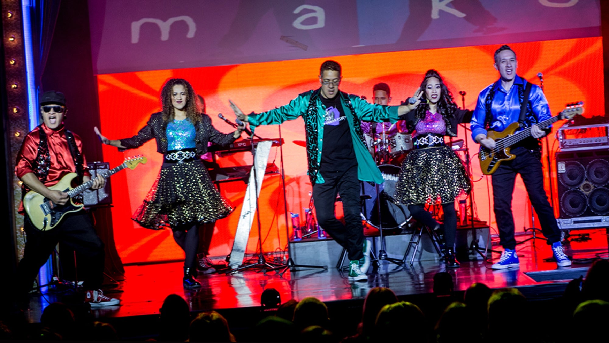 A band performs on stage with colorful outfits, electric guitars, a keyboard, and drums, in front of a red-lit background and an audience.