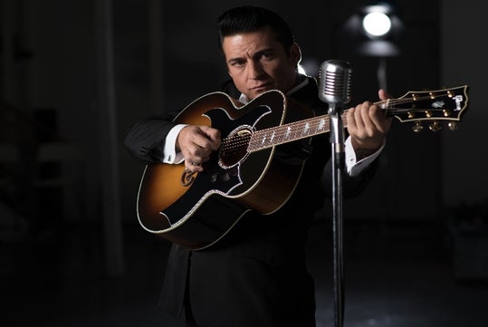 Shawn Barker in a black suit holding a guitar close to his upper chest in front of a microphone.