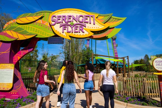 Five people walk toward the entrance of the Serengeti Flyer ride at an amusement park on a sunny day.