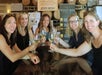 Five women sit around a table inside a winery, smiling and holding up glasses of white wine for a toast. Wooden barrels and promotional signs are visible in the background.